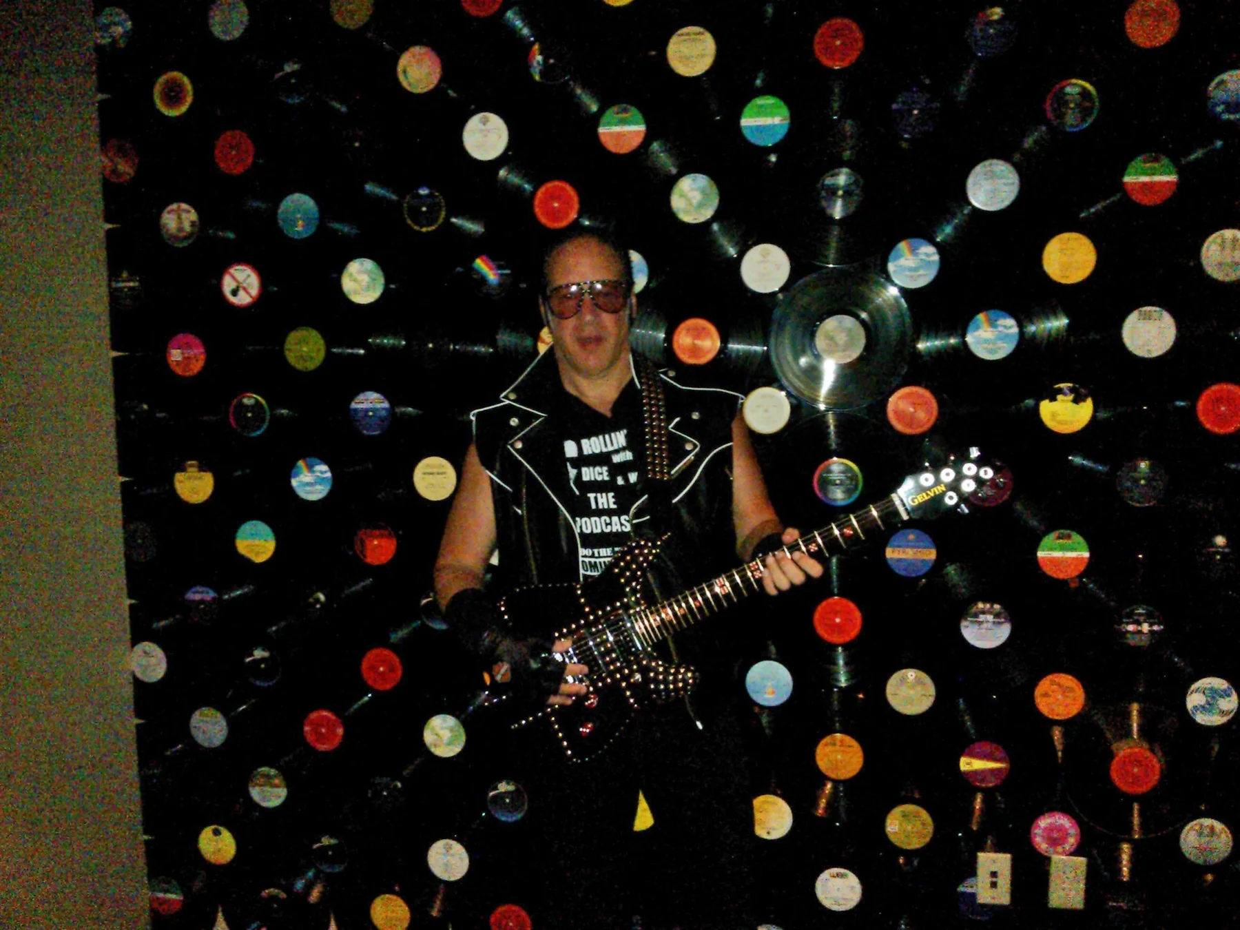 Andrew Dice Clay holding his custom Gelvin guitar in front of a wall of records